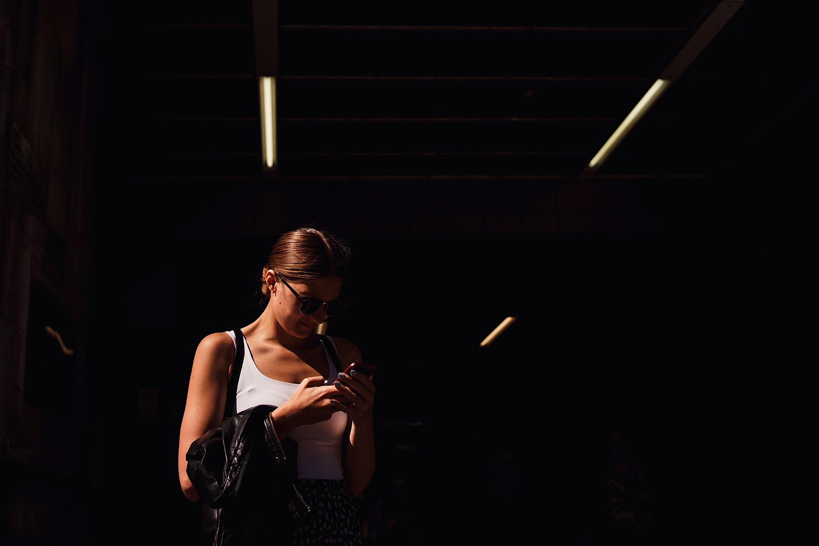 A girl on her phone with strips of light behind her at the entrance of Barbican station
