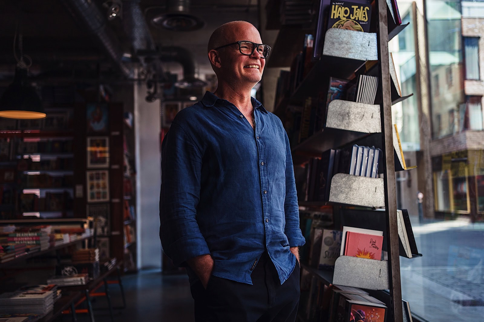 Commercial portrait of man wearing a blue shirt smiling, looking out the window of a bookshop