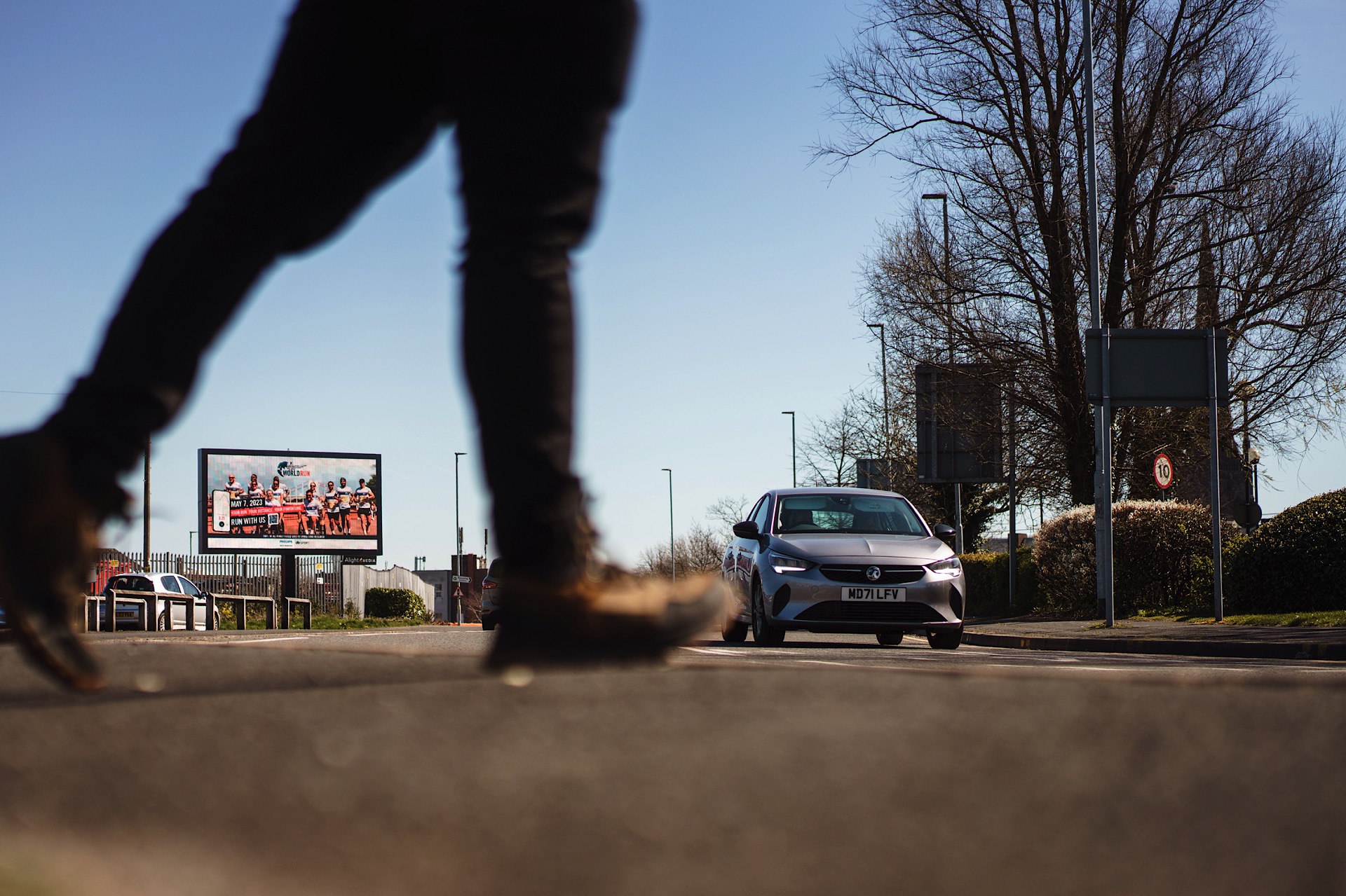 Person crossing a road as a car approaches. Trees and a billboard in the background on a sunny day.