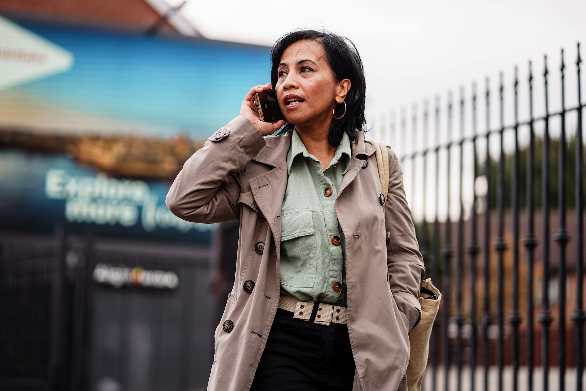 Person in a beige coat and green shirt talking on a phone, walking past a fence on a city street.