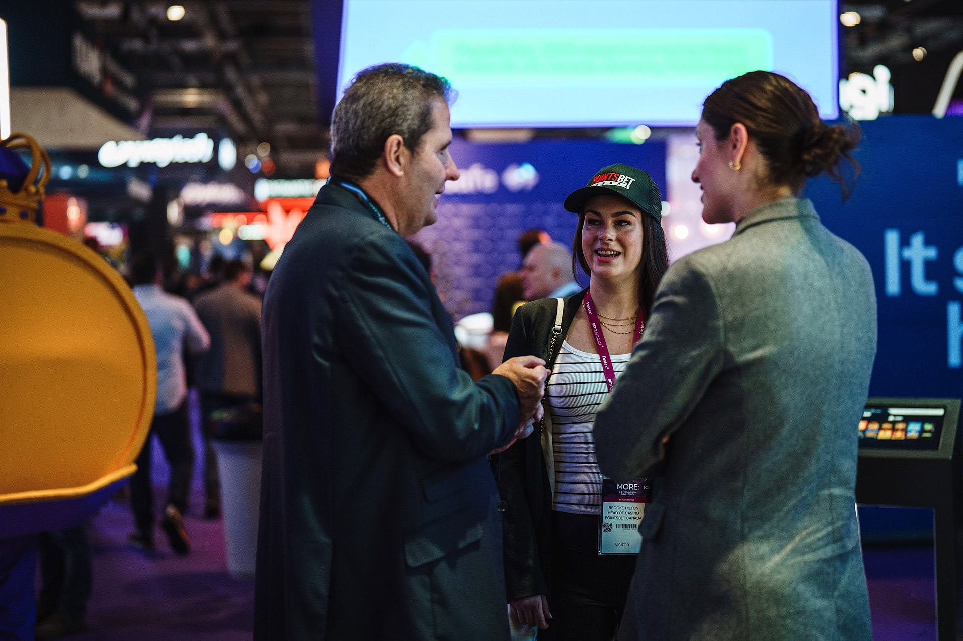 Three people are engaged in conversation at an indoor event with booths in the background. One person is wearing a cap and holding a bag, and another wears an overcoat.