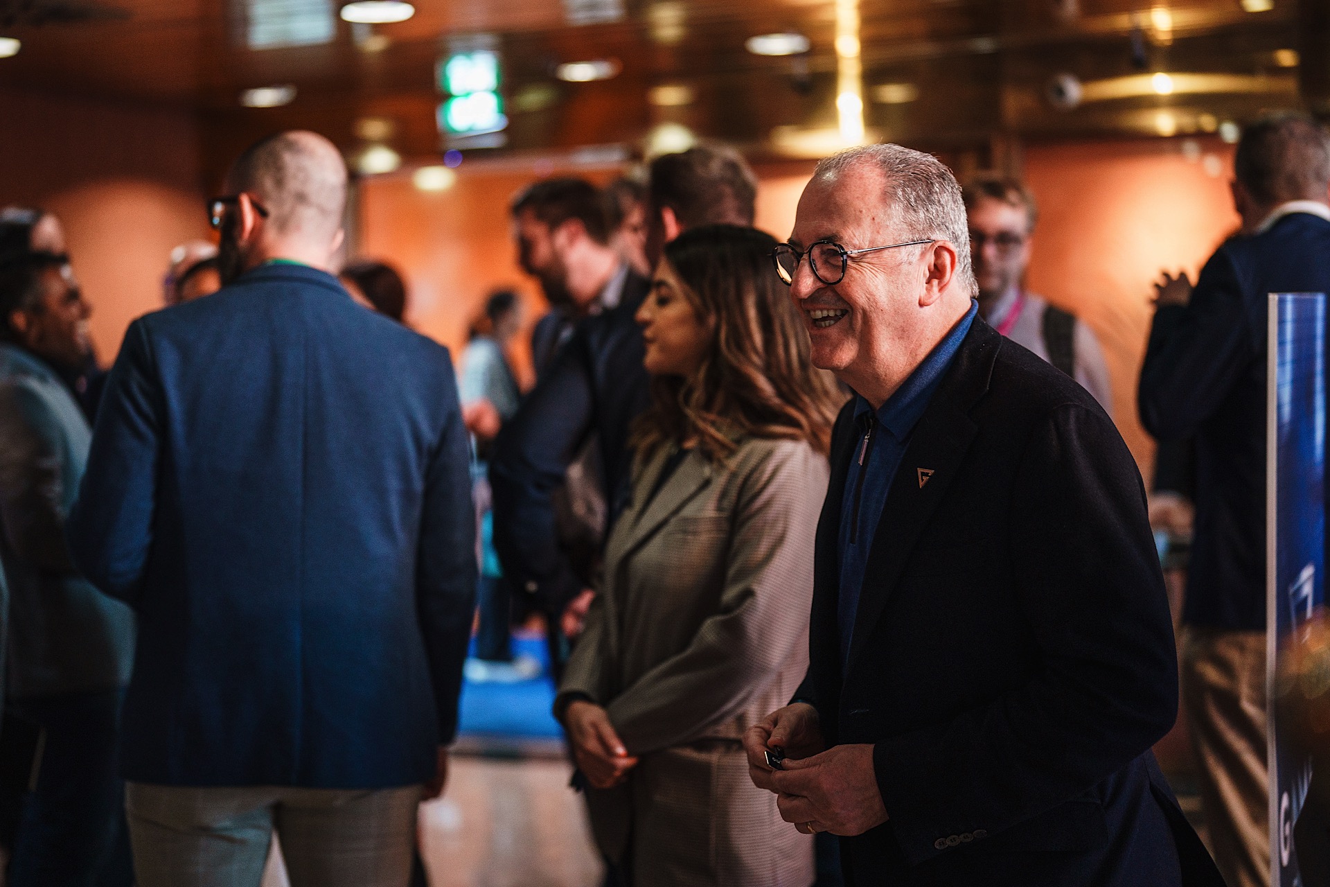A group of people in business attire are gathered indoors, with a smiling man in glasses and a dark suit in the foreground.