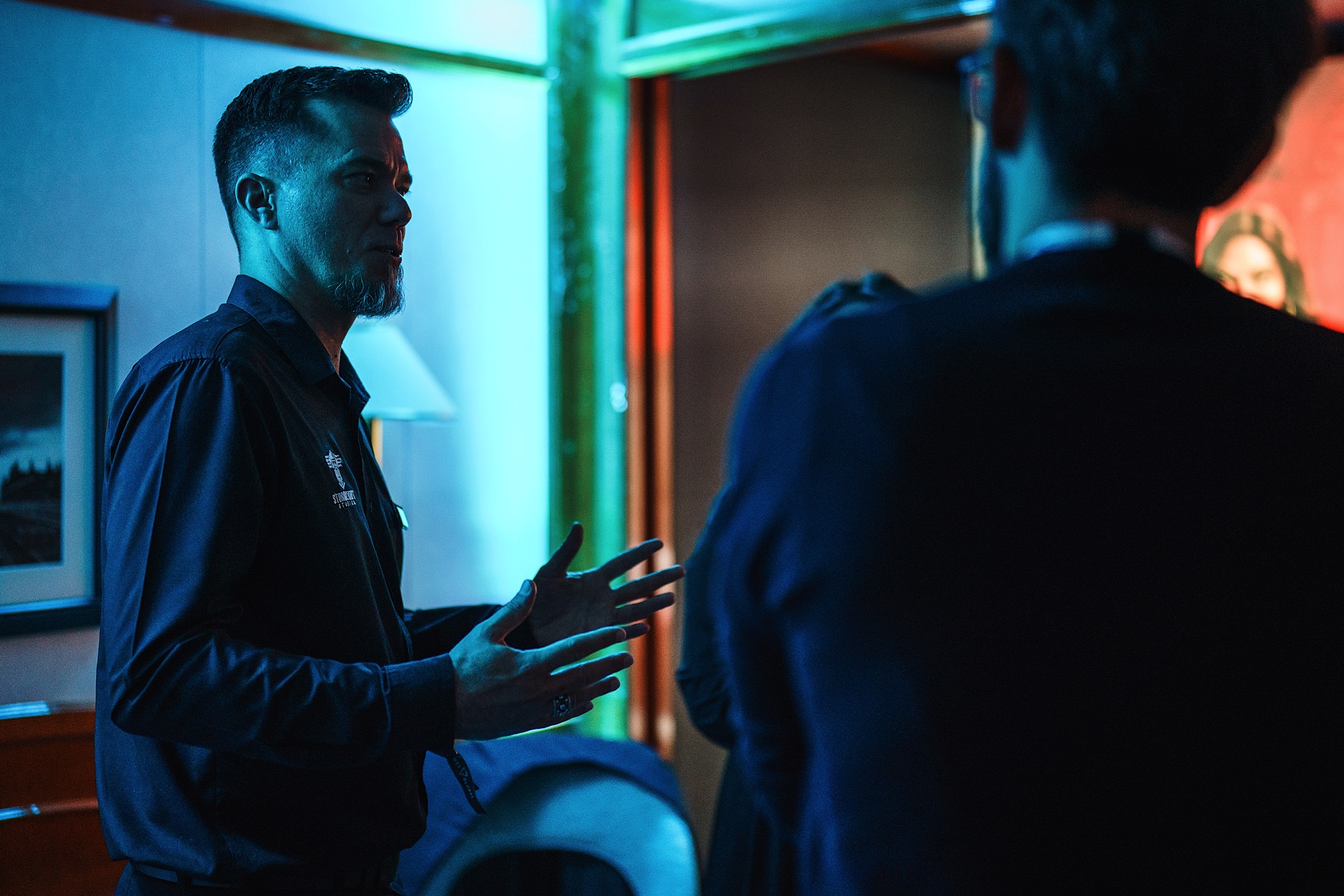 A man in a dark shirt gestures while speaking to two people in a dimly lit room with blue and red lighting.