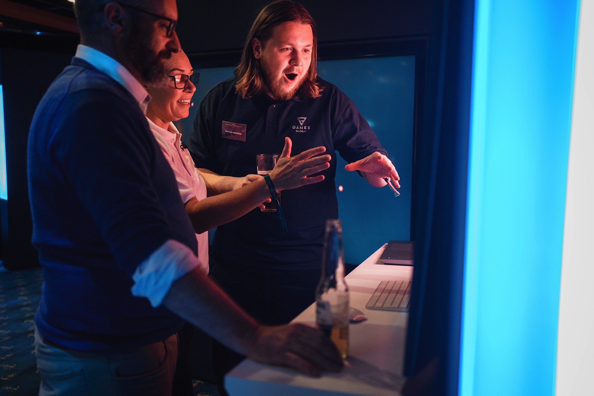Three people interacting with a touchscreen display in a dimly lit environment. One person gestures animatedly while others watch. A bottle sits on the counter nearby.