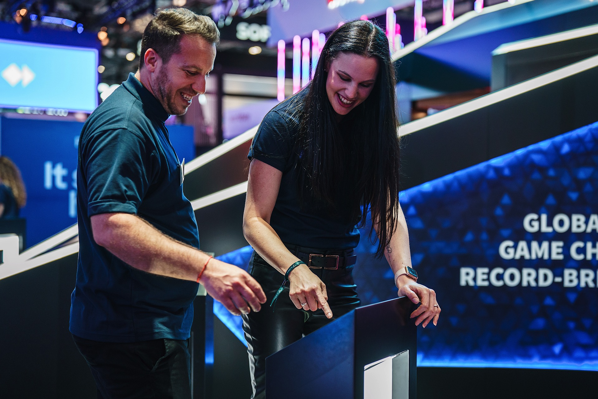 Two people in matching blue shirts happily interact with a digital kiosk at a tech-themed event, surrounded by blue and pink lighting.