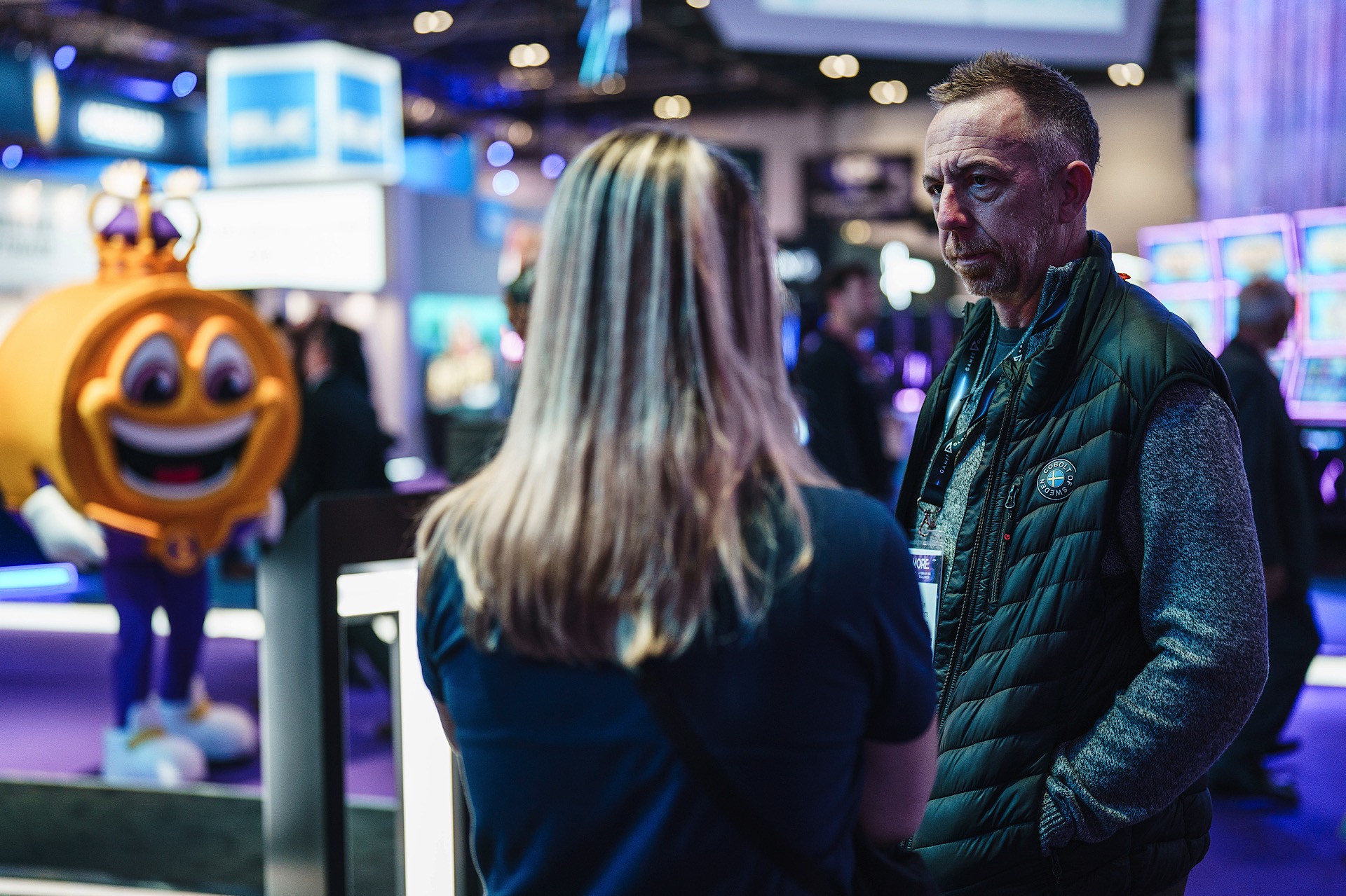 Two people in conversation at a busy indoor event, with a mascot in the background. The setting has blue and purple lighting.