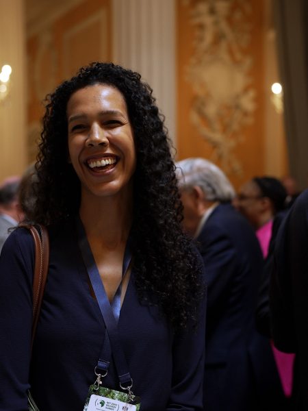A woman with long curly hair smiles in a crowded room. People in business attire are engaged in conversation. A chandelier and ornate decor are visible in the background.