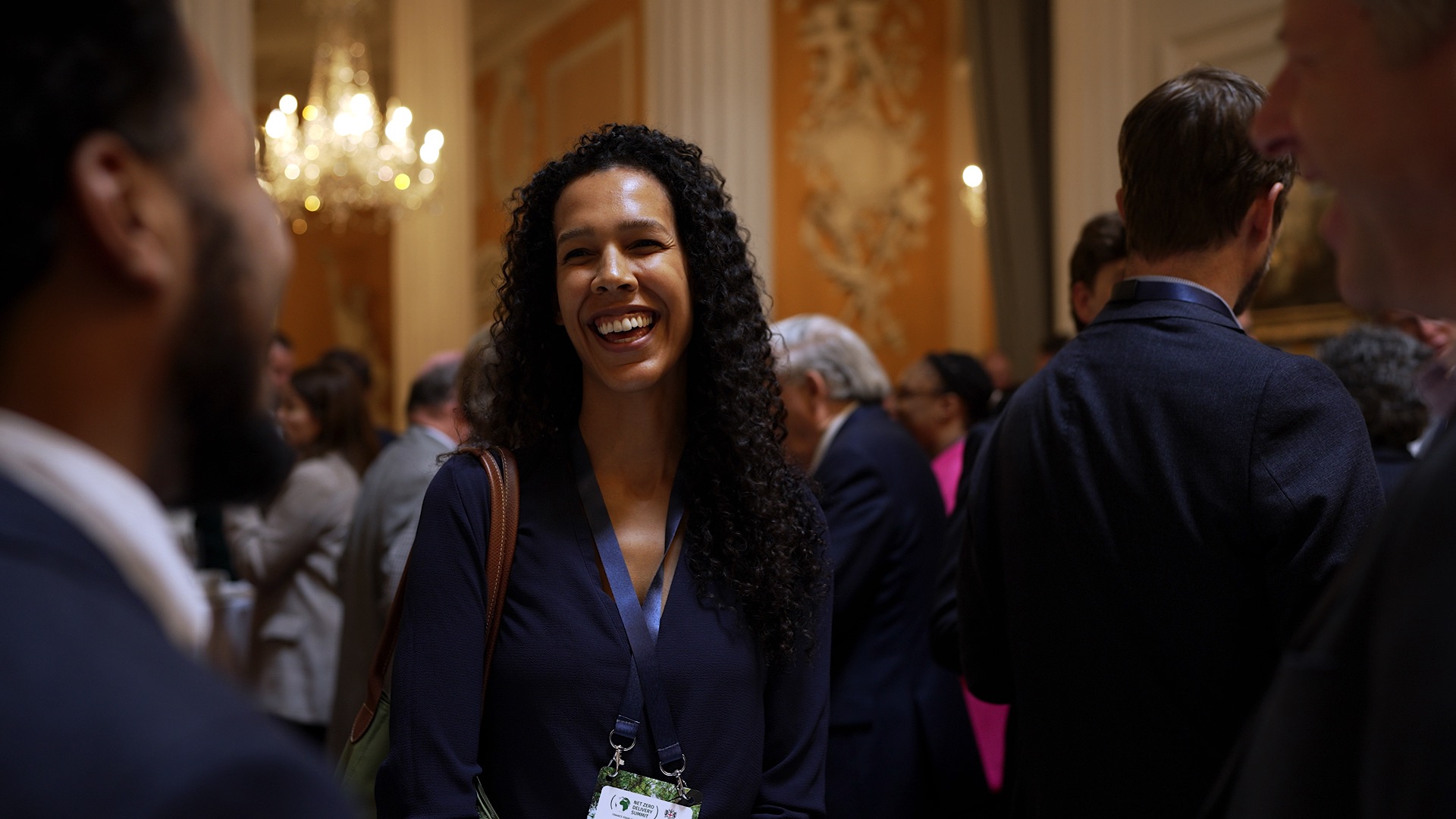 A woman with long curly hair smiles in a crowded room. People in business attire are engaged in conversation. A chandelier and ornate decor are visible in the background.