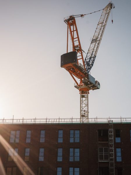 A construction crane towers over a brick building under a clear sky, with the sun shining bright.