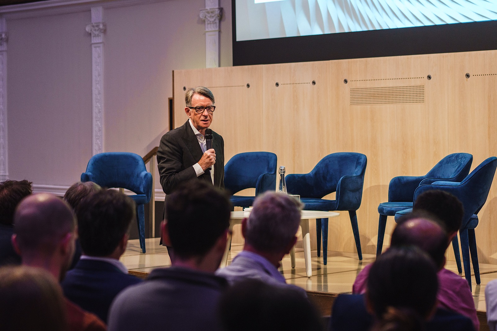 A man in a suit speaks into a microphone on stage before an audience, with several empty blue chairs behind him.