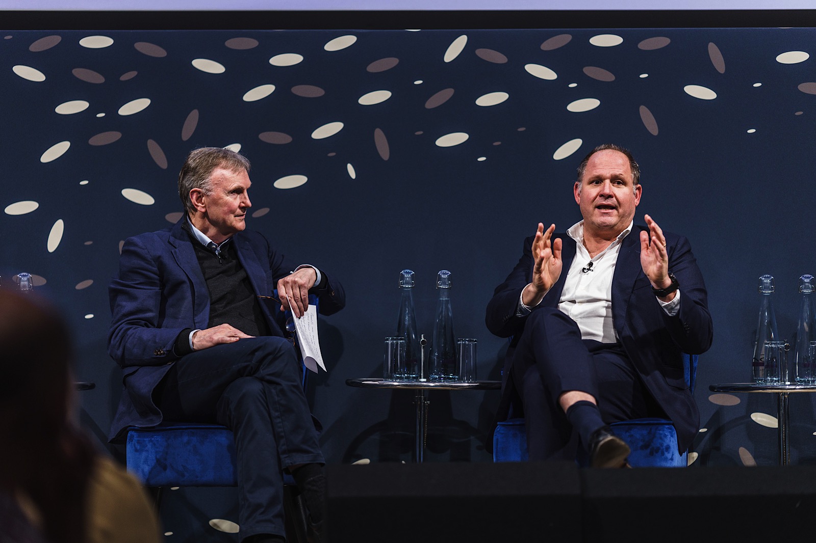 Two men in suits sit on stage in armchairs, engaged in discussion, with glass water bottles on small tables beside them and a patterned backdrop behind.