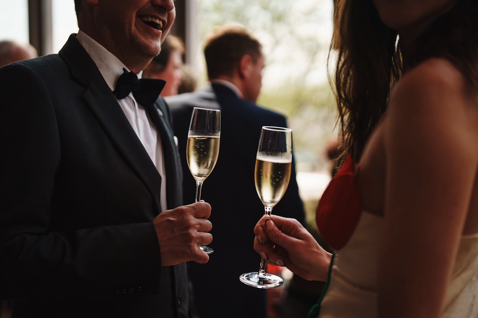 Two people dressed formally hold champagne glasses and converse at a social event, with other guests blurred in the background.