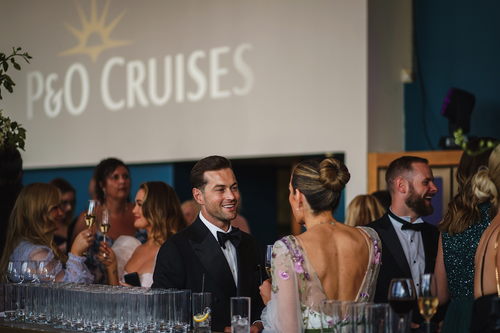Guests in formal attire socialize at a bar during a P&O Cruises event, with drinks and empty glasses visible in the foreground.