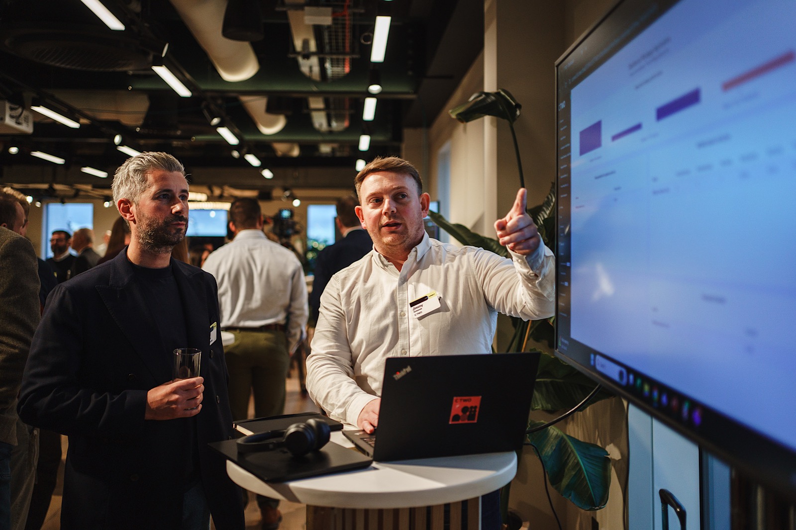 Two men stand beside a round table with a laptop, one pointing at a large screen displaying charts, in a modern office with people socializing in the background.