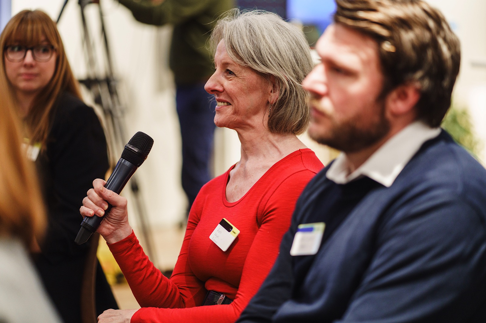 A woman in a red shirt holds a microphone and speaks during a meeting, with other attendees seated nearby.