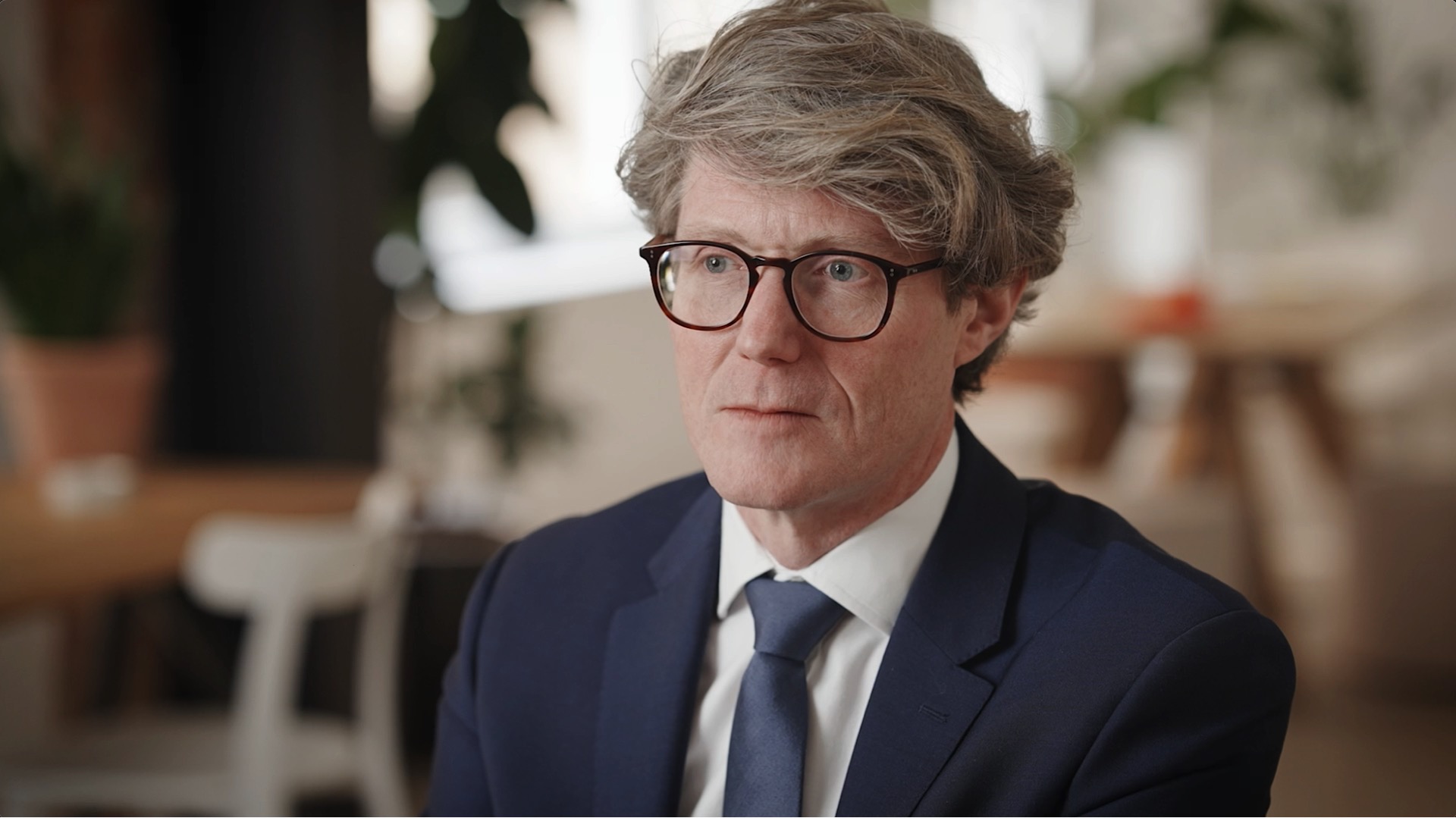 A man with gray hair and glasses wearing a navy suit and tie sits indoors, looking forward with a neutral expression.