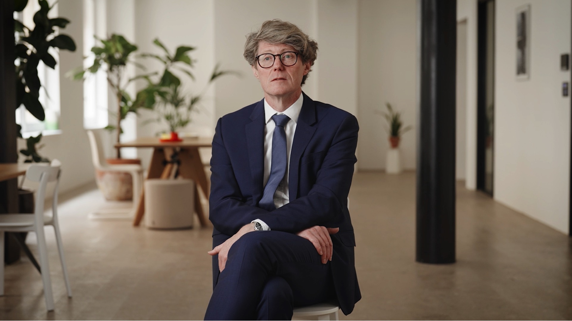 A man in a blue suit and glasses sits with legs crossed on a chair in a modern, well-lit room with plants and minimalist decor.