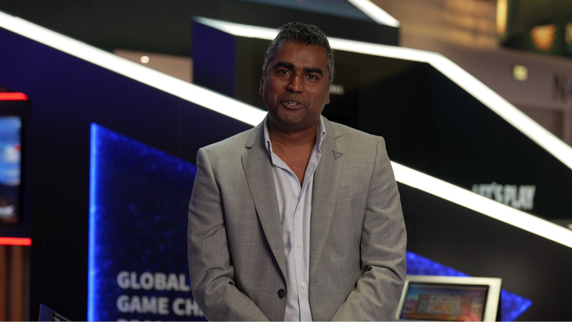 A man in a light gray suit stands in front of a modern, angular exhibition booth with digital displays and geometric lighting.