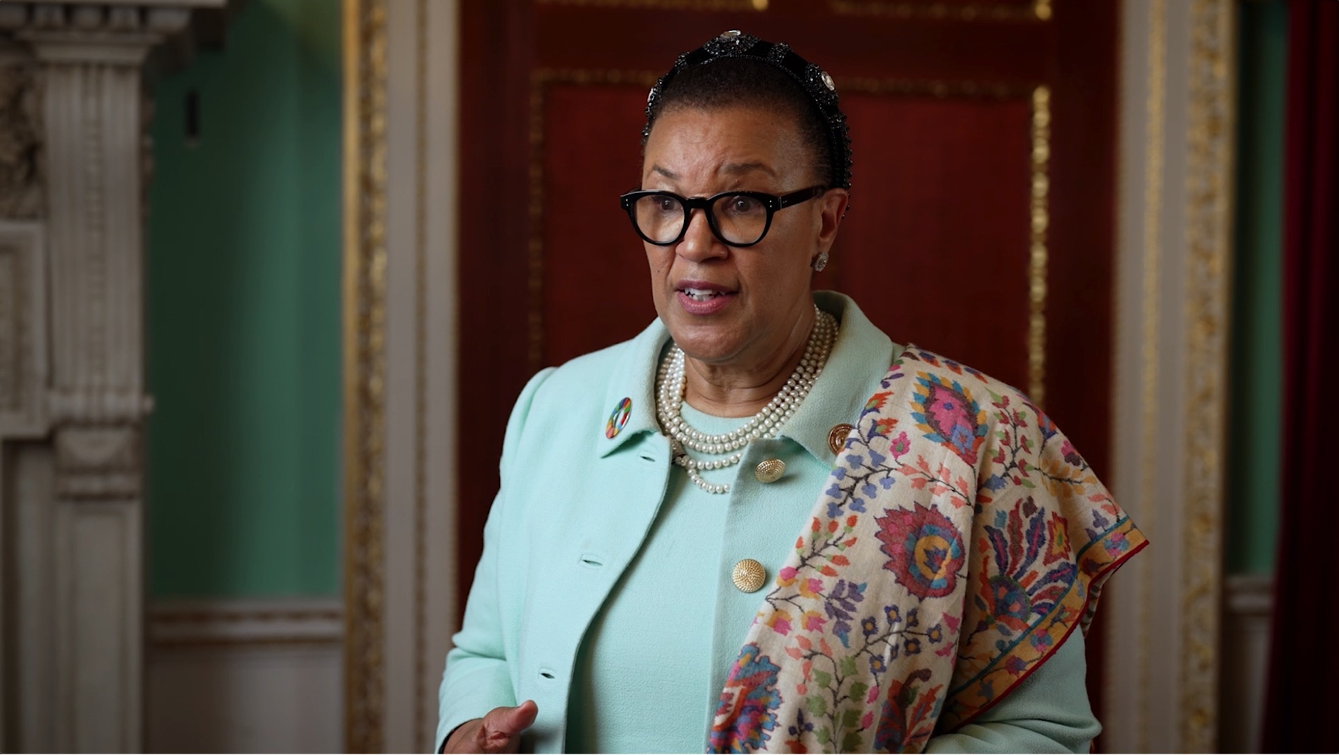 A woman wearing glasses, a light green jacket, and a patterned shawl stands indoors in front of a decorative wall, looking ahead and speaking.