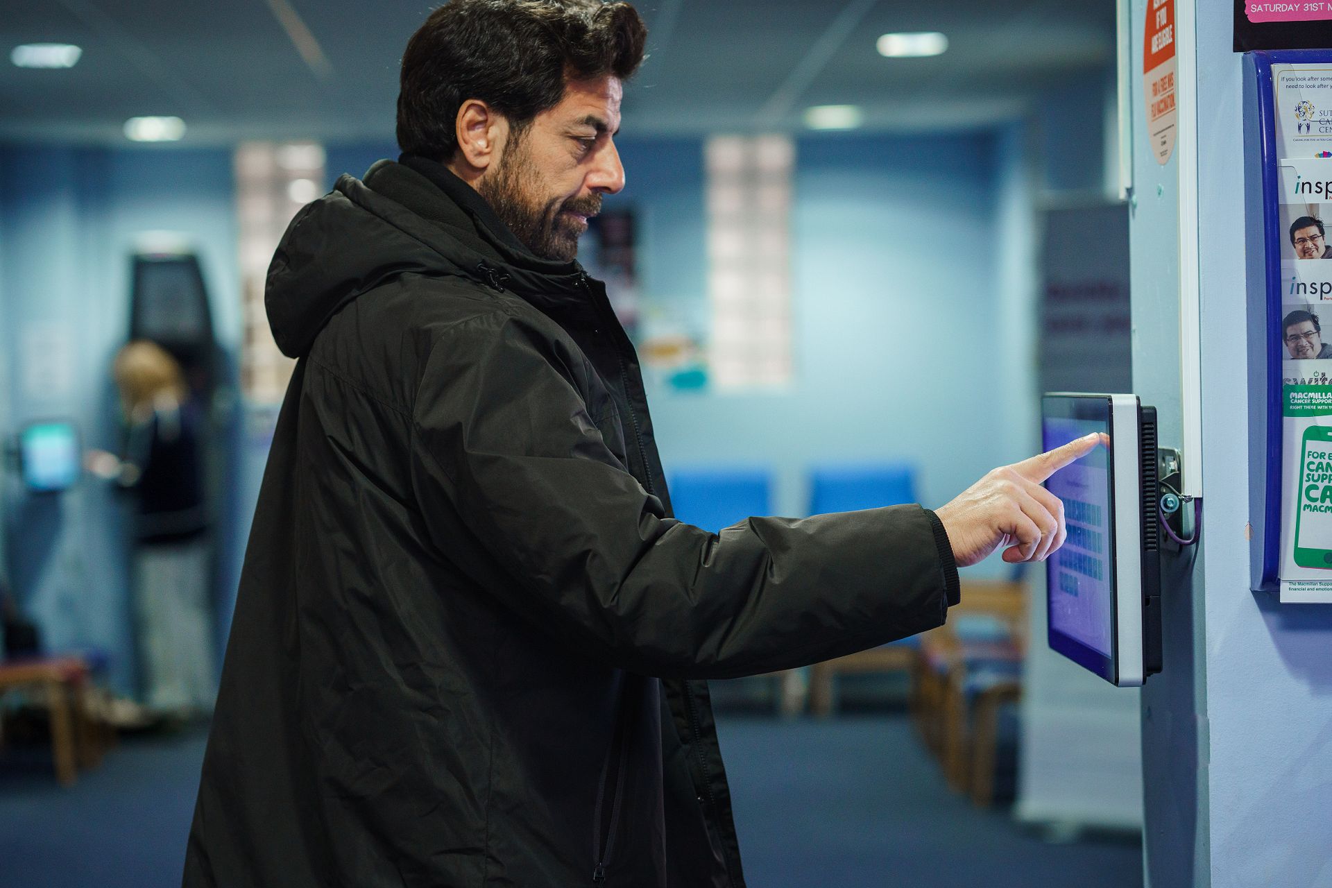 A man in a dark coat uses a touchscreen check-in kiosk in a waiting area with blue chairs and posters on the wall.